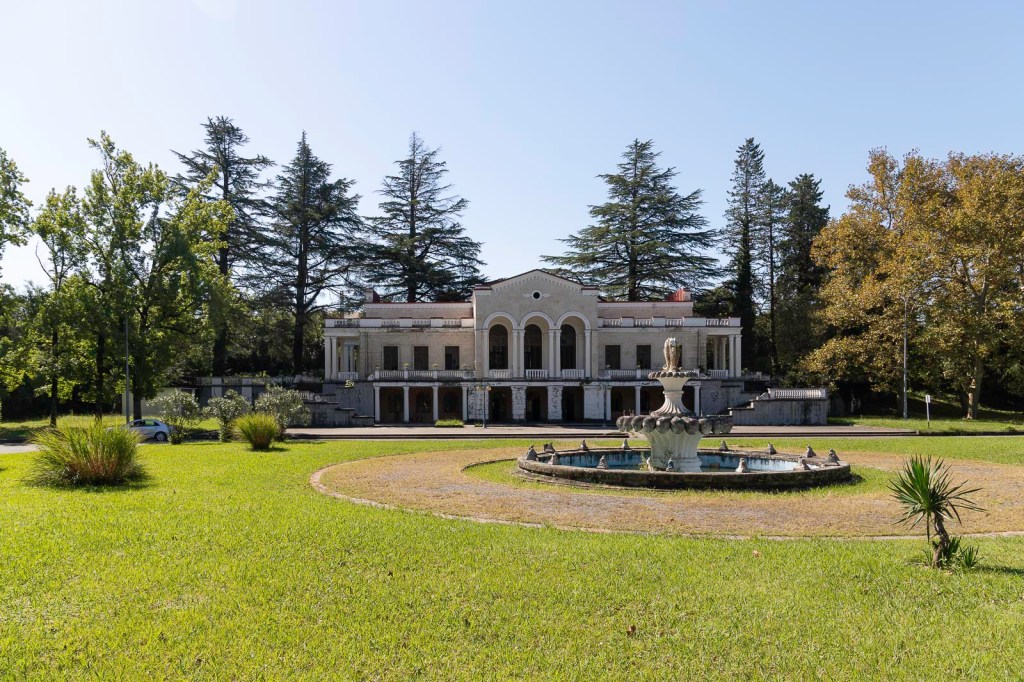 Een groot, historisch gebouw met een fontein op de voorgrond, omgeven door groene gazons en hoge bomen.