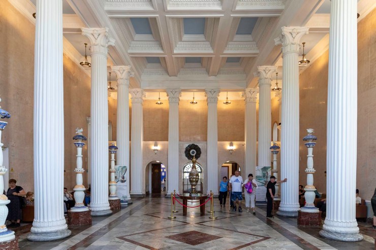 Interieur van een grote hal met hoge zuilen, marmeren vloeren en een decoratief plafond. Mensen bewegen zich door de ruimte, terwijl een opvallend object in het midden staat, omringd door een rode afzetting.