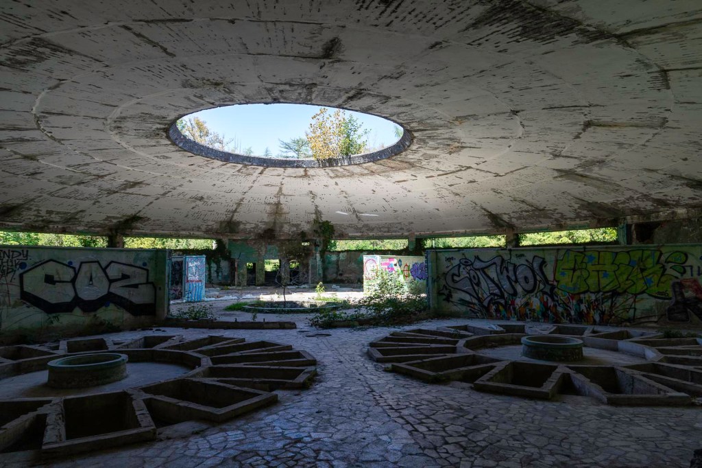 Een vervallen ruimte met een ronde opening in het plafond, omringd door muren met graffiti en vegetatie die door de vloer groeit.