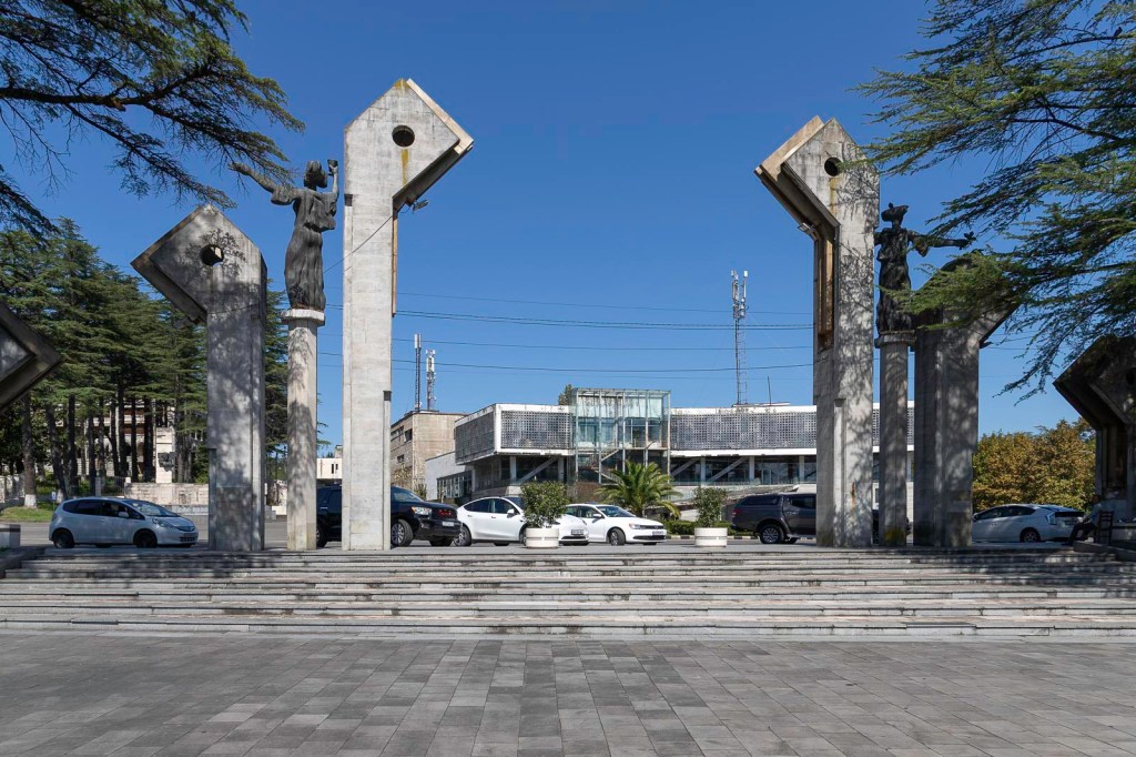 Een open ruimte met grote betonnen monumenten in de vorm van pijlen, met standbeelden aan de zijkanten; op de achtergrond is een modern gebouw zichtbaar en er staan verschillende auto's geparkeerd.