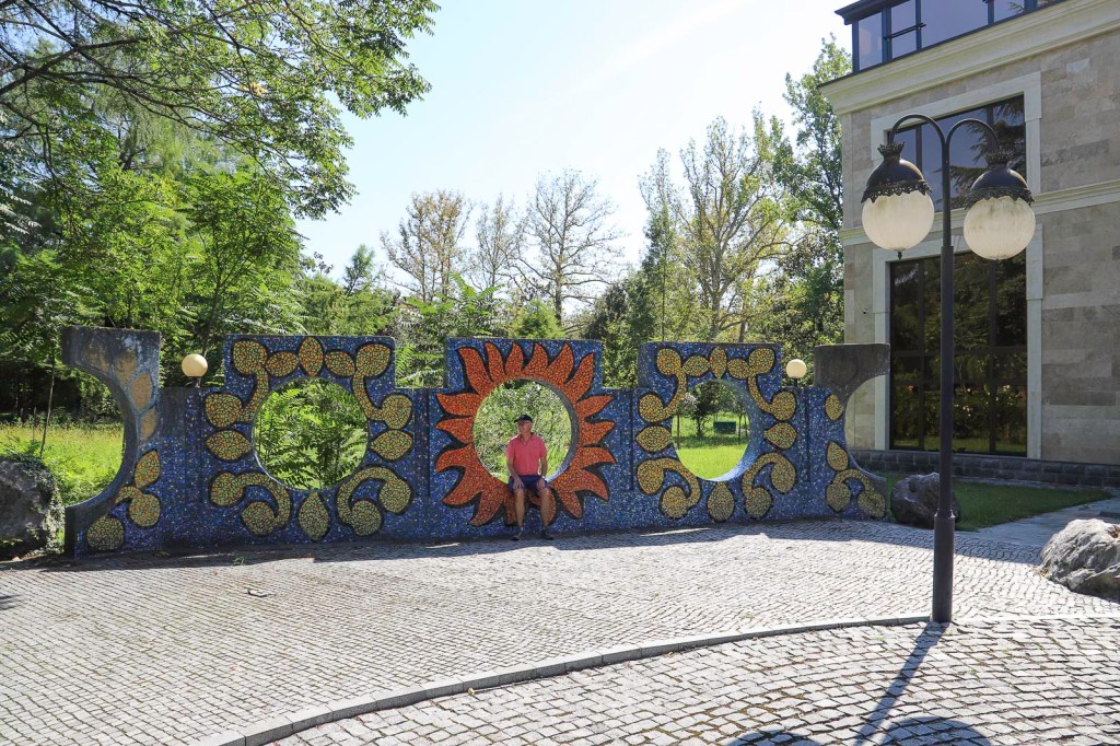 Een kleurige mozaïekmuur met verschillende patronen, waaronder een grote zon en cirkels, staat in een groene tuin. Een persoon in een roze shirt zit op de muur, terwijl moderne gebouwen op de achtergrond zichtbaar zijn.