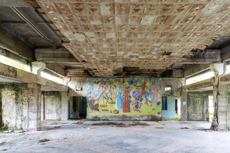 Een vervallen interieur met een beschadigd plafond en een kleurrijk muurschildering op de achtergrond.