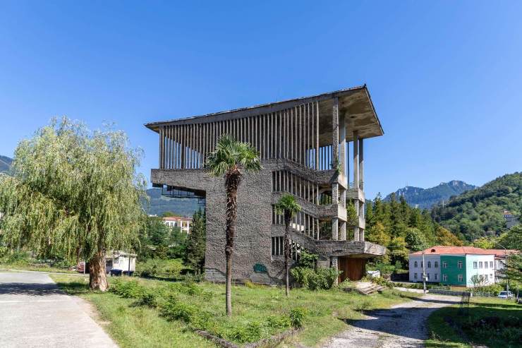 Een modern en onvoltooid betongebouw in een groene omgeving met bomen en bergen op de achtergrond.