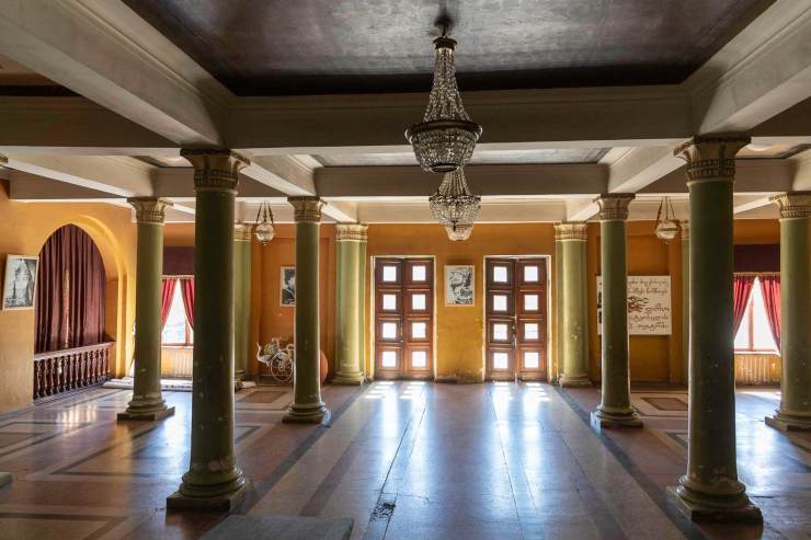 Interieur van een grote zaal met kroonluchters en zuilen, met vensters en gordijnen in een warme kleurstelling.