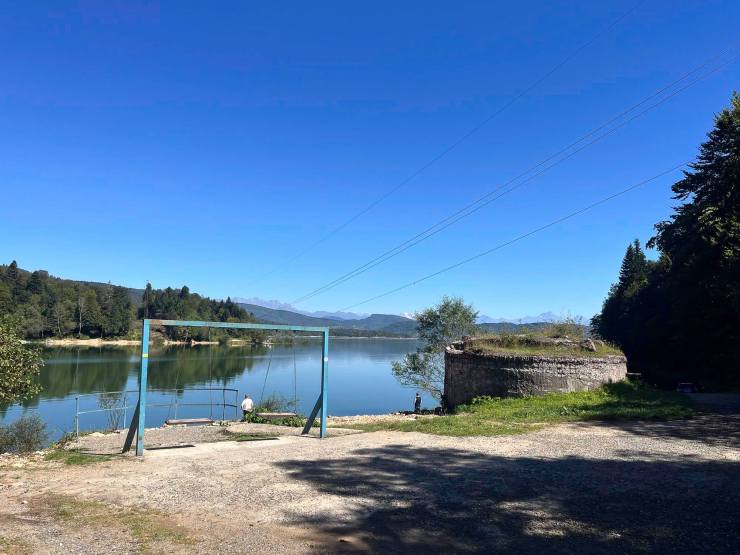Uitzicht op een rustige rivier met aan de oever een schommel en een oude stenen structuur, omgeven door bomen en bergen onder een heldere blauwe lucht.