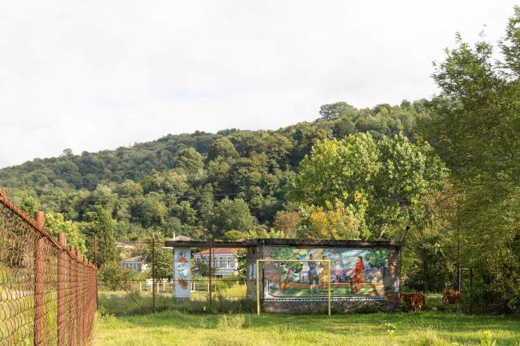Een vervallen schuur met muurschilderingen staat op een groene weide, omgeven door bomen en een heuvel op de achtergrond.