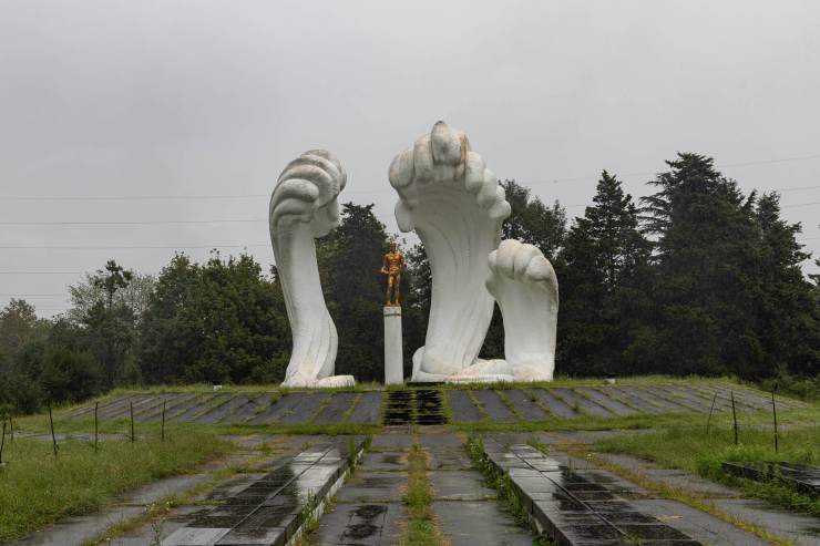 Beeld van een sculptuur met grote witte handen die een standbeeld van een man omringen, in een groene omgeving met bomen op een regenachtige dag.