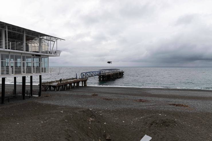 Een verlaten pier boven het water met een kustlijn op de achtergrond, onder een bewolkte lucht.