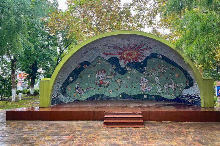 Een amphitheater met een kleurrijke mozaïekwand, versierd met natuurbeelden zoals bloemen, zonnen en figuren die verschillende activiteiten uitvoeren, omgeven door bomen.