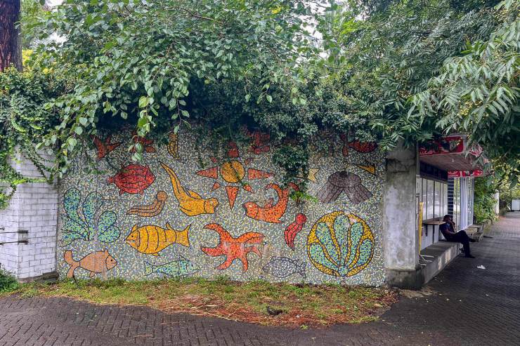 Mosaicwand met kleurrijke zeedieren, waaronder vissen en schelpen, omringd door groene bladeren en een halte met een persoon die zit.