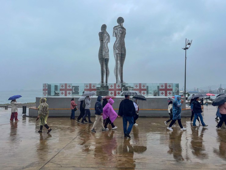Mensen wandelen onder paraplu's langs een moderne sculptuur van twee figuren op een natte boulevard, met regenachtige lucht op de achtergrond.